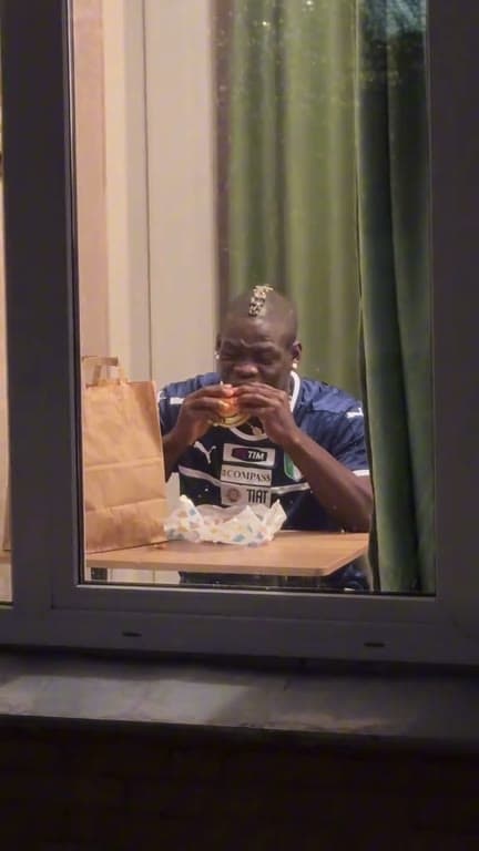 A person eating a burger looks toward the camera from a window at night.